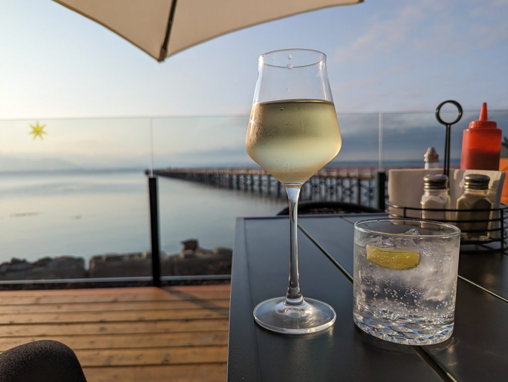 A close up of two drinks on a table with a condiment basket, with a glass balcony overlooking the ocean and long pier.