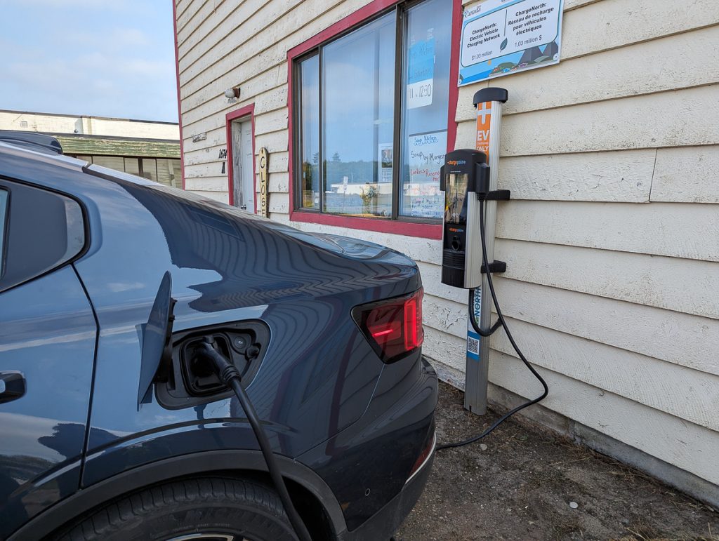 The rear of an electric car connected to a charger on a creamy wooden building.