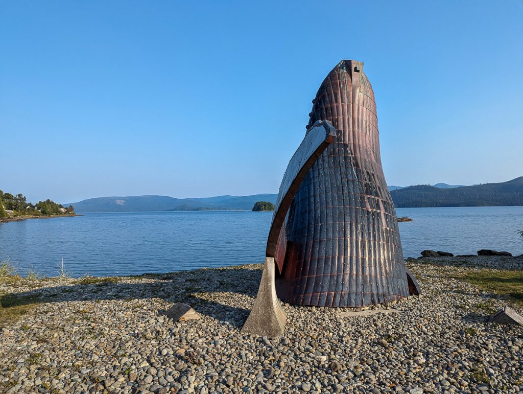 A sculpture of a whale breaching from a pebbled ground with water and forested hills in the background.