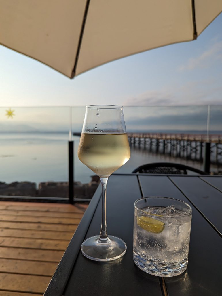 A close up photo of a glass of white wine next to a glass of clear spirits on a table looking towards a blurred background of a wharf leading into water.