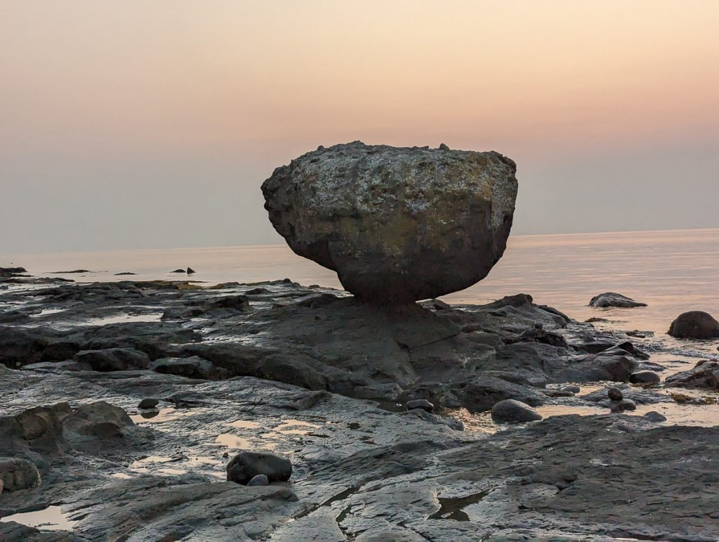 A large rock balancing on top of a rocky shoreline next to water with a very light pink sky behind.