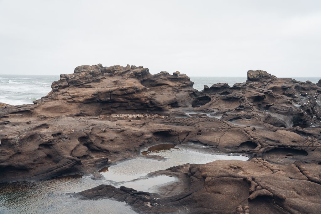 Dark brown undulating rocks and rockpools next to water on an overcast day.