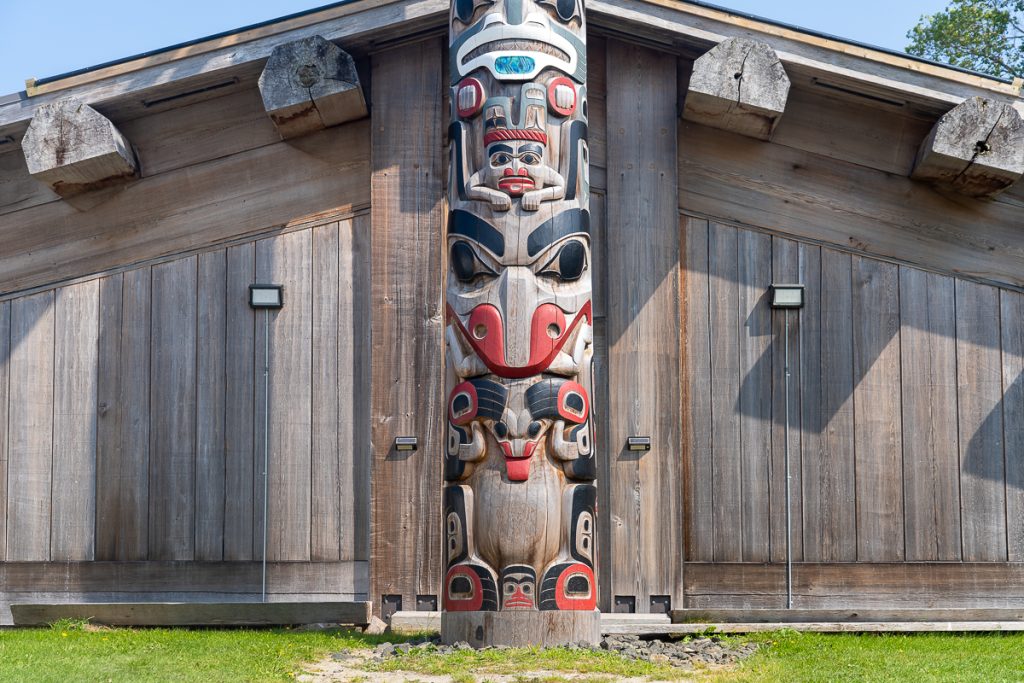 A wooden monumental pole with creatures carved and painted standing in front of a wooden building.