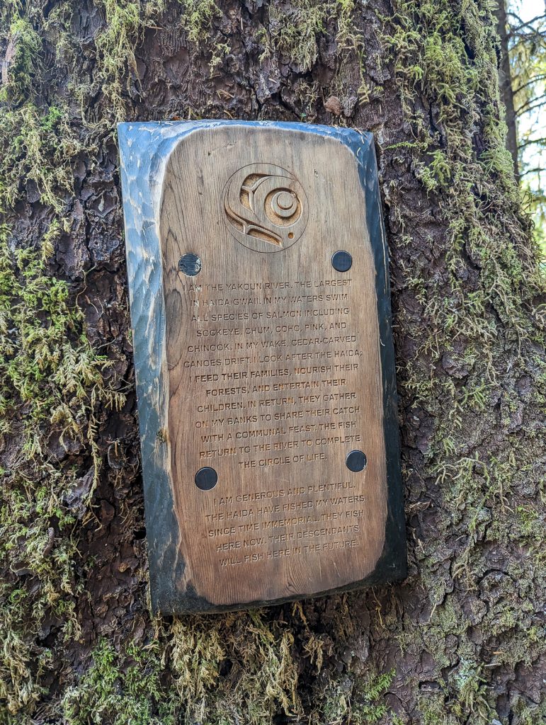 A carved wooden plaque attached to a tree trunk that tells the story of the Yakoun River in Haida Gwaii.