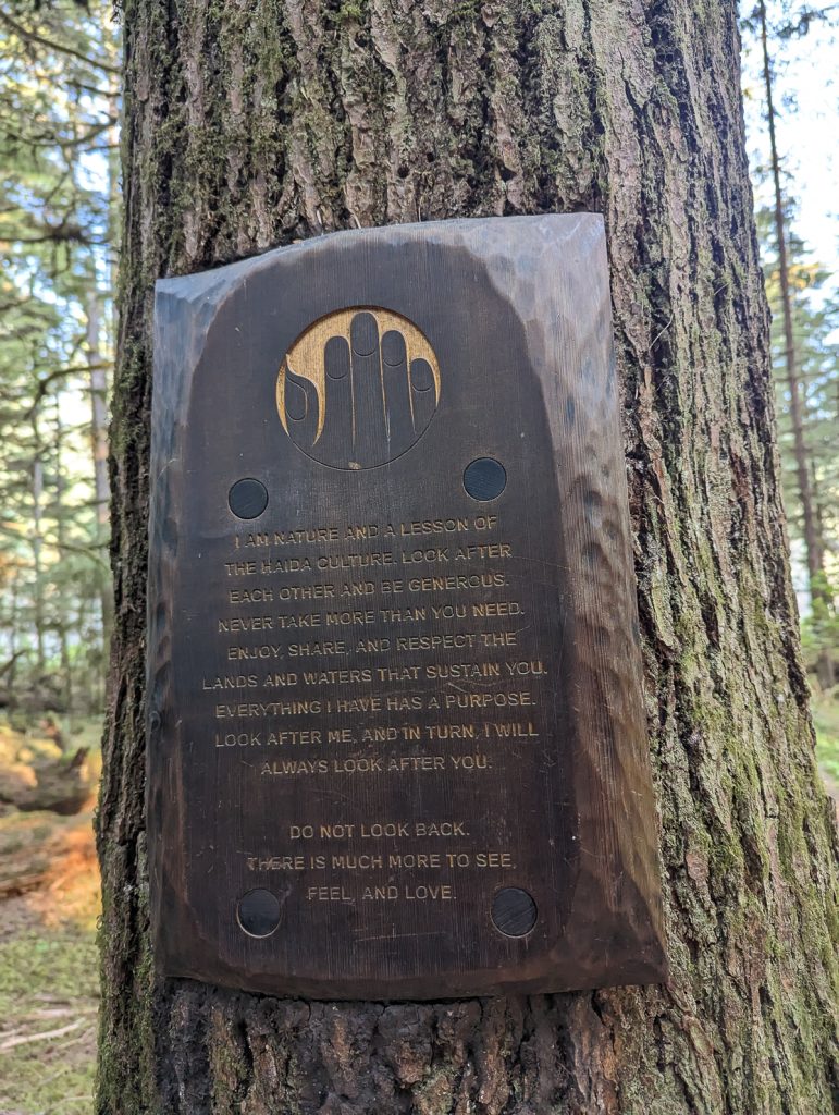 A carved wooden plaque attached to a tree trunk in a forest with text telling a story about Haida culture.