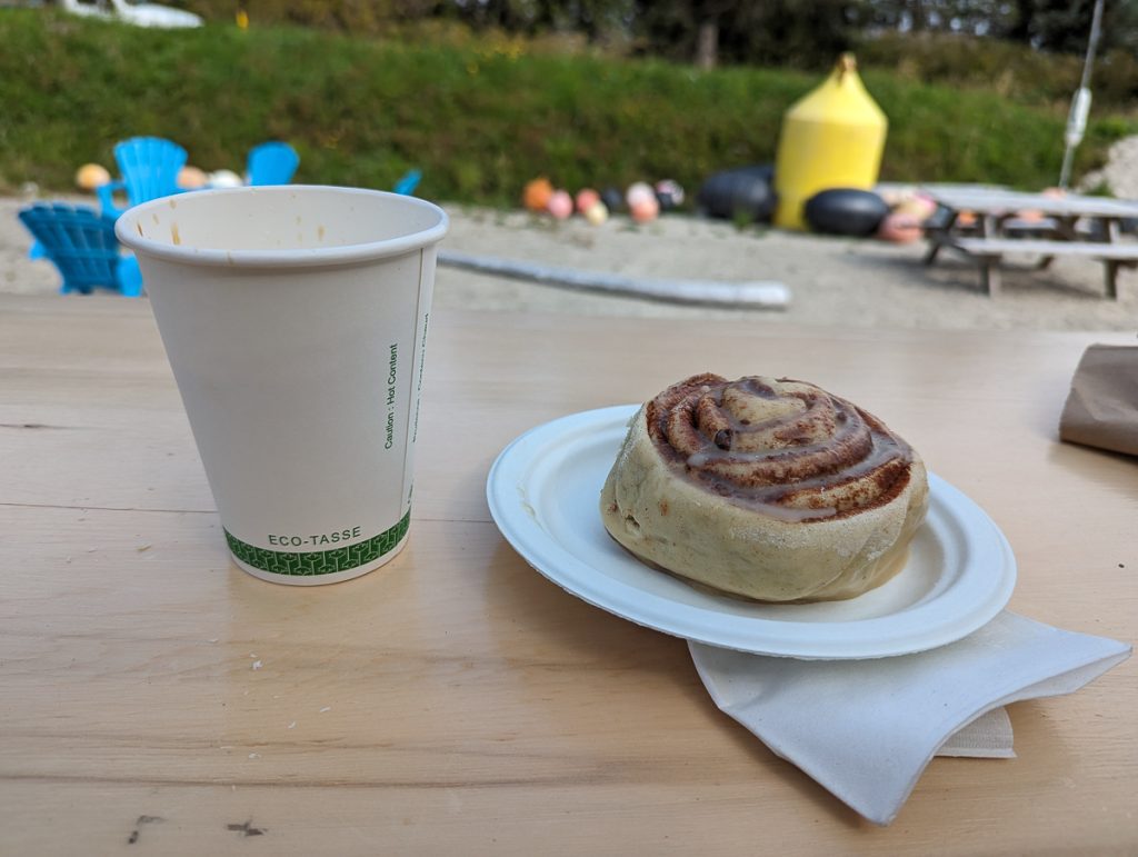 A close-up shot of a paper cup and a cinnamon roll on a paper plate on a wooden surface with sand and chairs in the background.