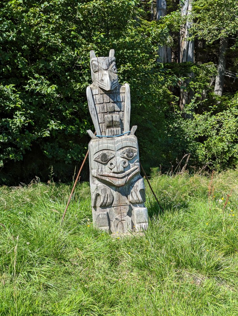 An old Haida carving stands weather-worn in the grass on Haida Gwaii