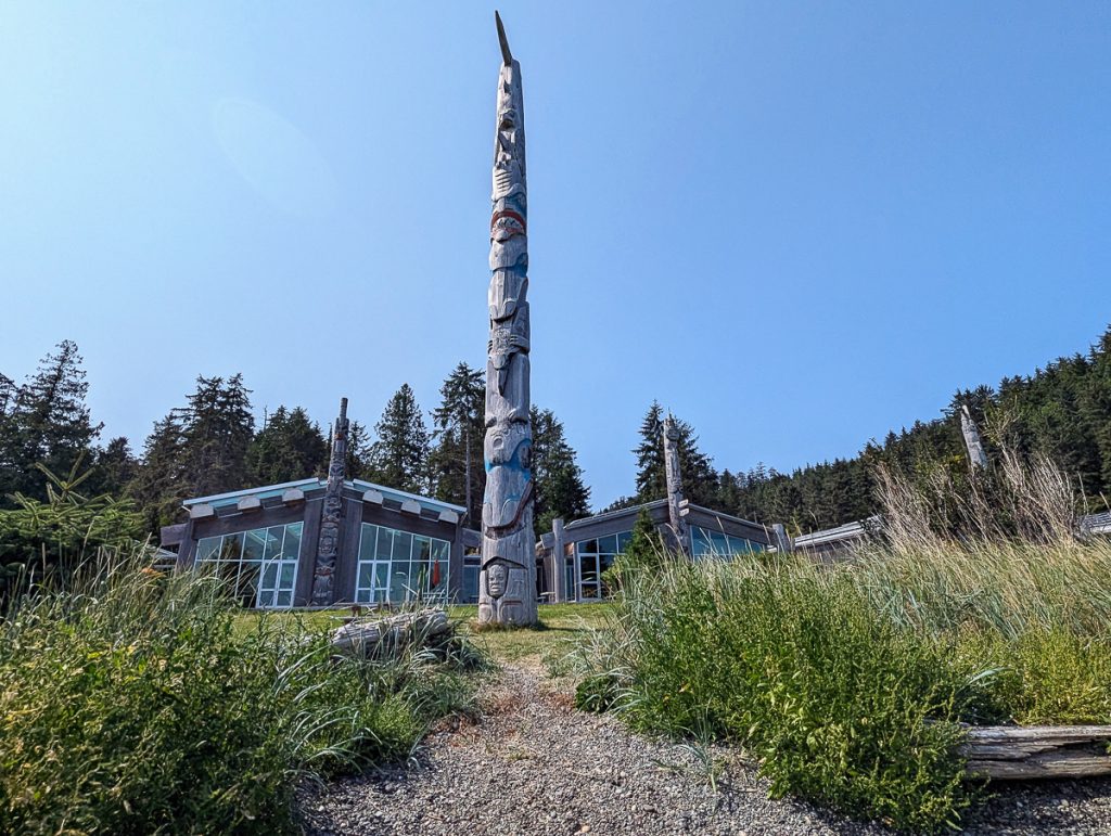 A carved wooden monument pile rising from grass with a building and more poles in the background.
