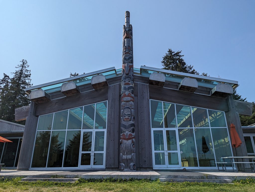 A wooden building with large glass windows and a carved wooden monument pole in front with blue sky behind.