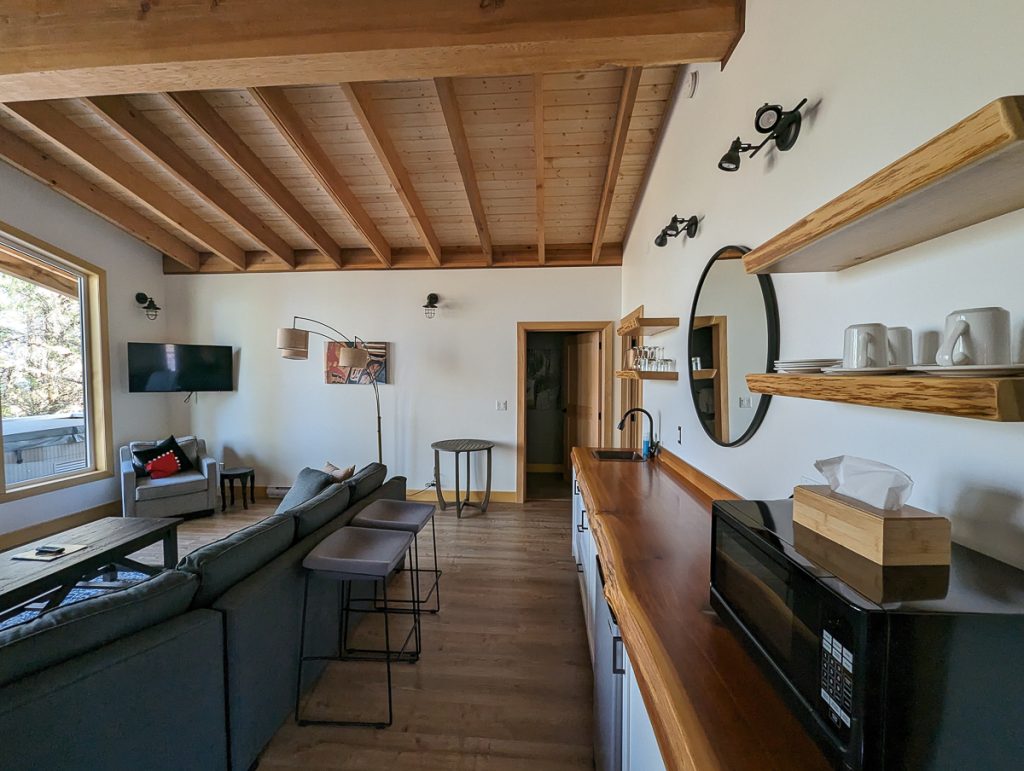 A room with white walls and exposed wooden beams containing a large couch with coffee table and tv on the wall, with a wooden kitchen bench against the wall.