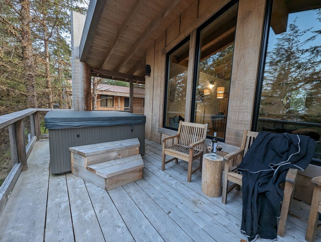 A large covered wooden balcony with a covered hot tub, chairs and a log side table holding a bottle of wine and glasses.