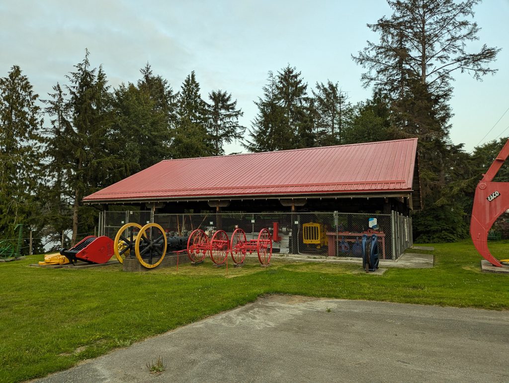 Antique agricultural equpment lined up on grass in front of a covered area with a red roof.
