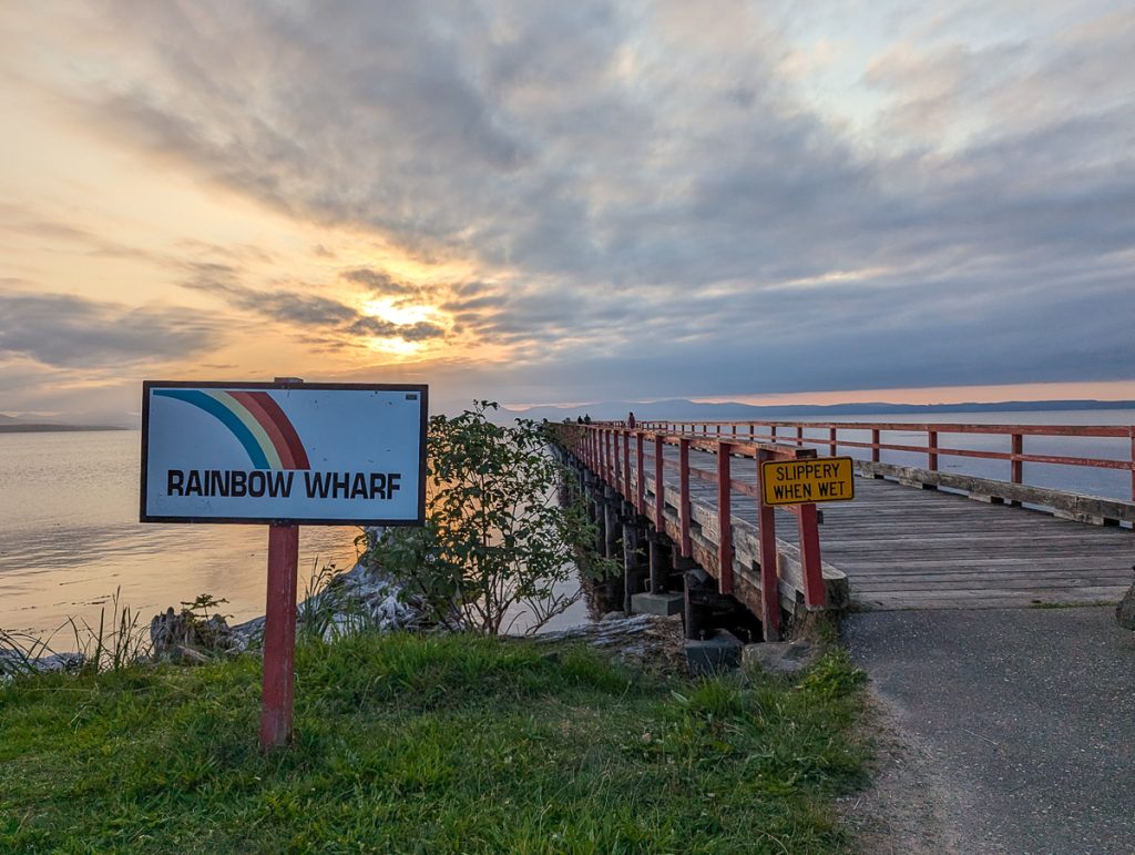 A wharf next to water at sunset with a sign saying "slippery when wet" and another sign with a rainbow painted on it with the words "Rainbow Wharf."