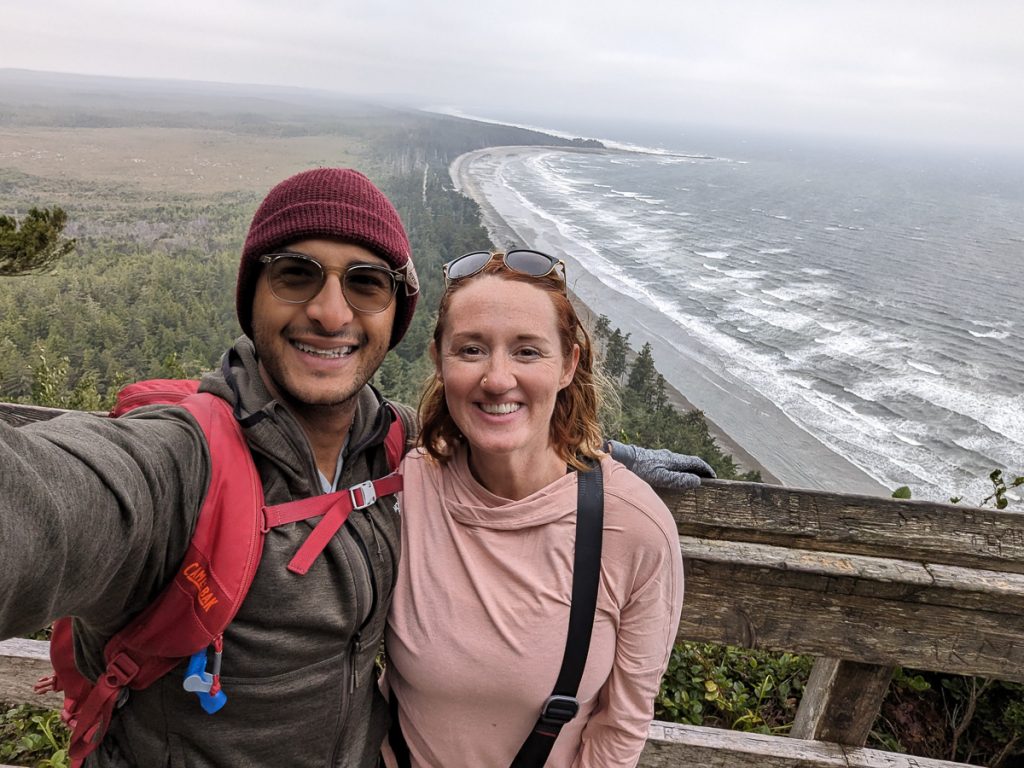 A man and woman smiling and taking a selfie at a viewpoint with woods and a beach behind them.