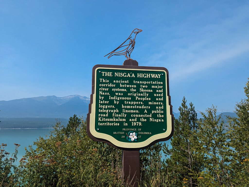 A green highway information sign with a raven sculpture on top and a lake and mountains in the background.