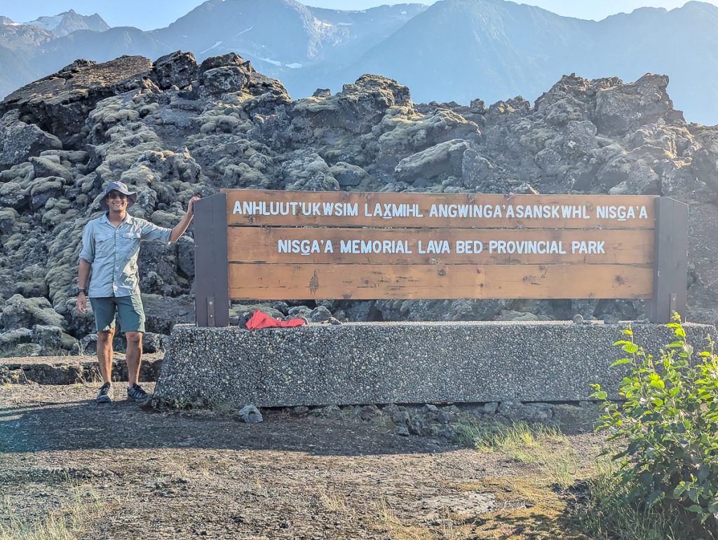 A man standing in front of rocks with his hand on a wooden sign that says "Anhluut'ukwsim Lax̱mihl Angwinga'asanskwhl Nisg̱a'a, Nisga'a Memorial Lava Bed Provincial Park."