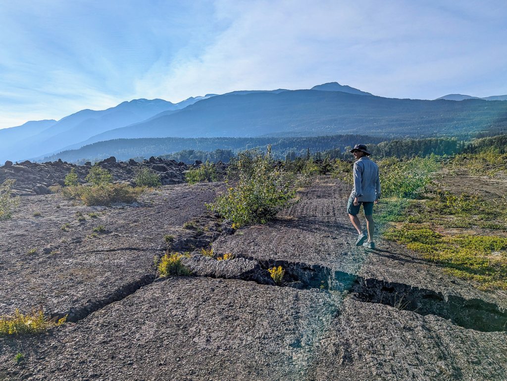 A man walking across a cracked lava rock plain with mountains in the distant background.
