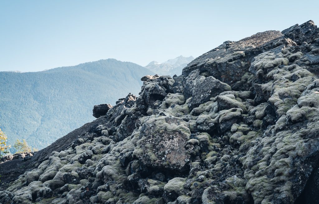 A rocky landscape of moss-covered lava rocks with mountains and blue sky in the background.