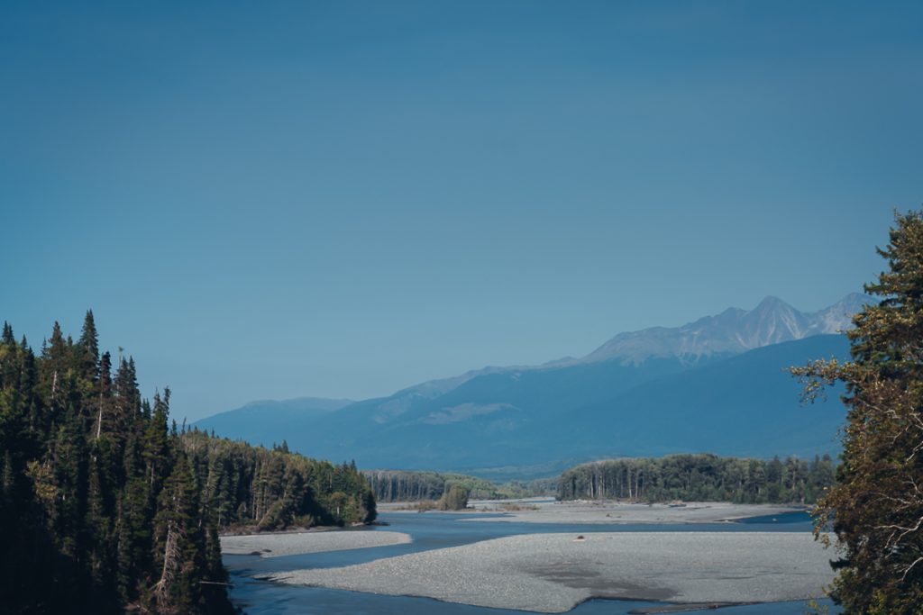 A river with rocky outcroppings into it, surrounded by pine trees and mountains in the distance with blue skies above.
