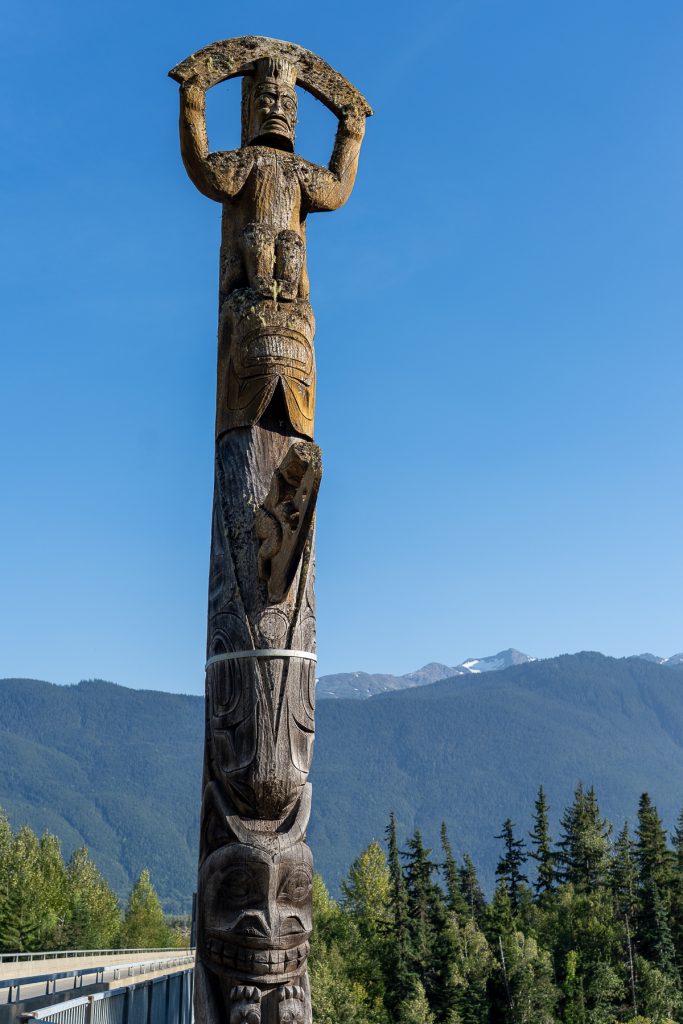 A Nisga'a Nation Orca Clan totem pole next to a road with mountains in the background.
