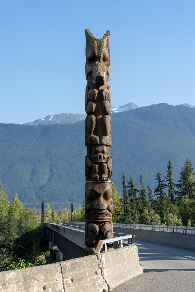 A Nisga'a National Wolf Clan totem pole next to a road with mountains in the background.
