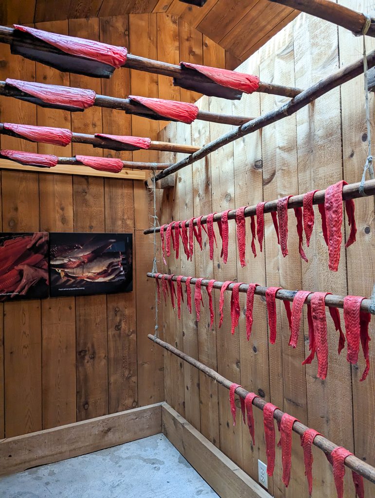 The interior of a traditional Nisga'a Nation smokehouse with red fish hanging over wooden poles.