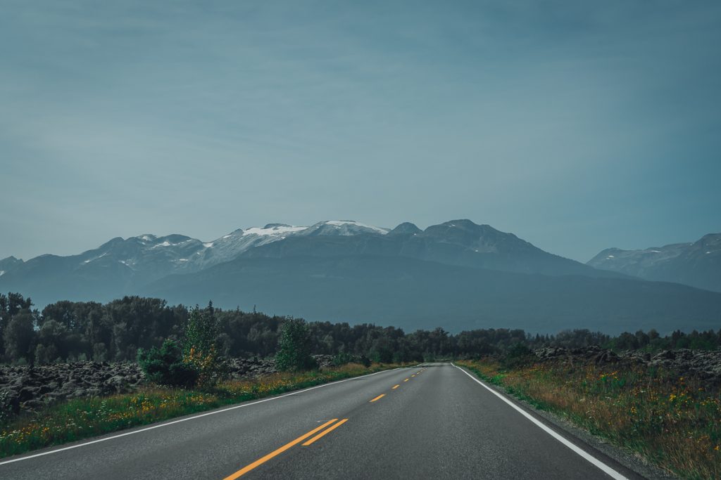 An empty road with greenery on either side and mountains capped with snow in the background.