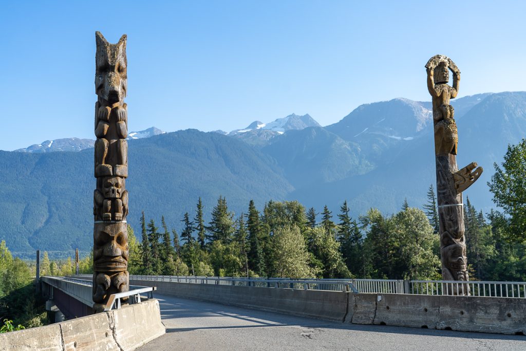 Two wooden totem poles carved with animals on either side of a road bridge, with mountains in the background.