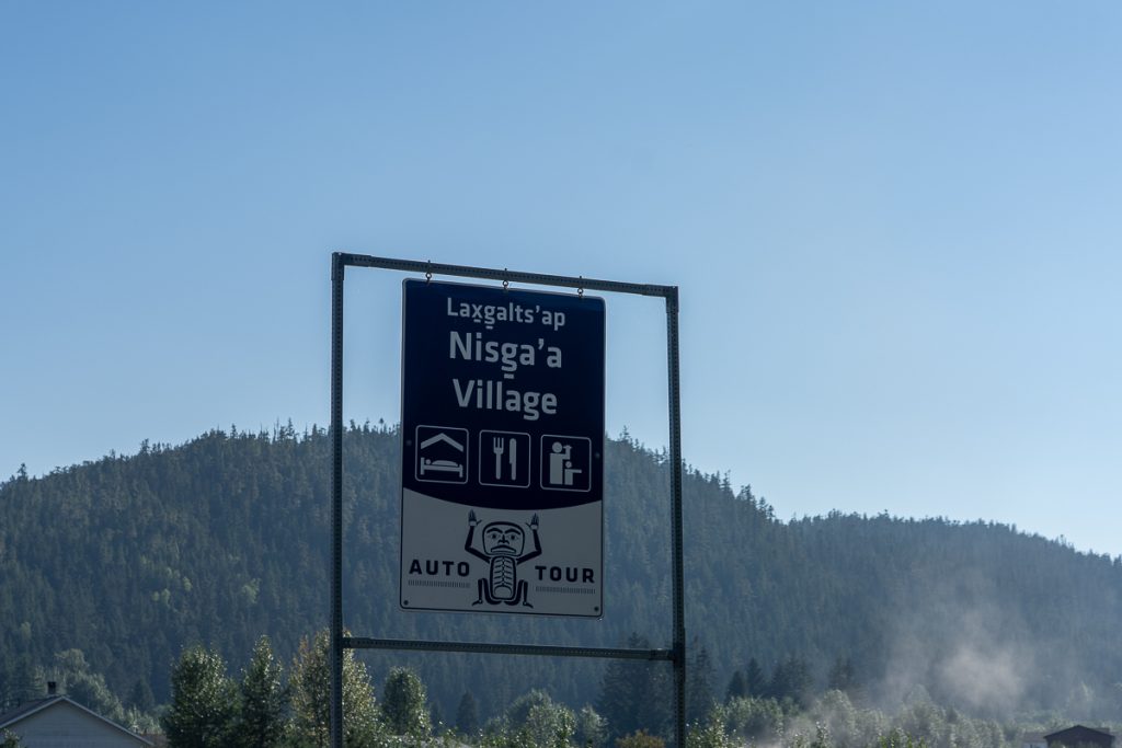 A roadside sign with text saying "Laxgalts'ap Nisga'a Village Auto Tour" and symbols for accommodation, dining and people.