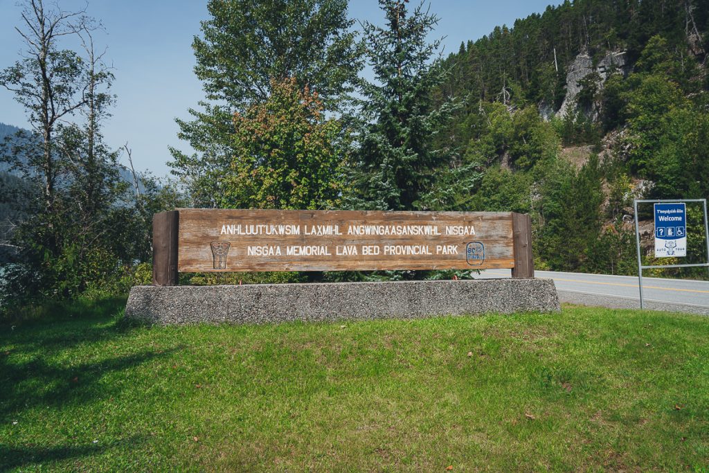 A roadside sign of wood on metal with text saying "Nisga'a Memorial Lava Bed Provincial Park" in English and the local language.