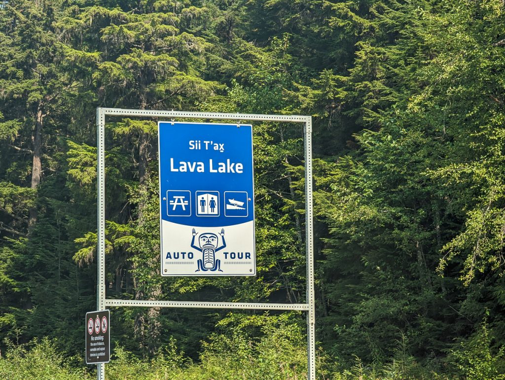 A blue roadside sign saying "Sii Táx Lava Lake Auto Tour" with symbols for picnic benches, toilets and a boat ramp.