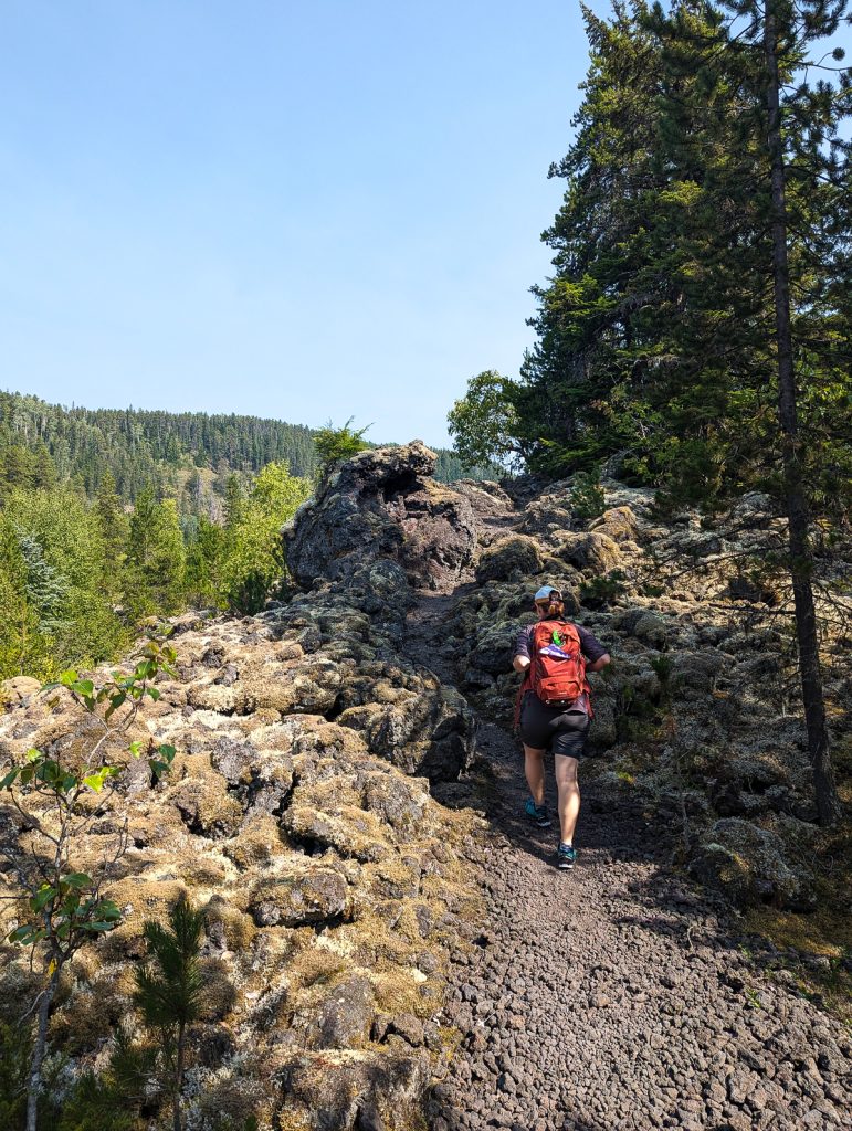 A person wearing a backpack seen from behind as they walk through a rocky landscape with trees and blue skies ahead of them.