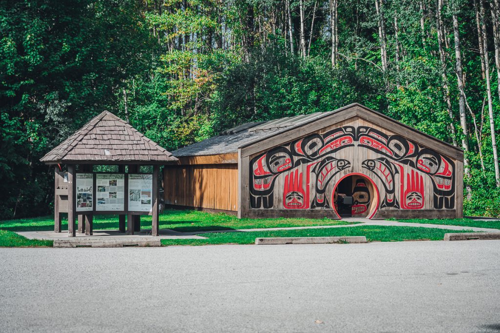A wooden shelter with informational signs next to a wooden building painted with red and black animal symbols and green trees behind.