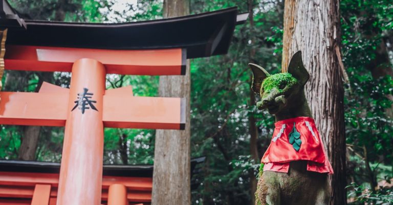 A Japanese Fushimi Inari shrine with a moss-covered statue of an animal in front and trees behind.