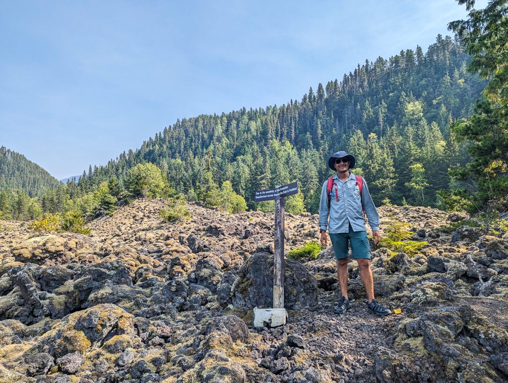 A man in hiking gear standing on rocky ground next to a signpost with a forested hill behind him.
