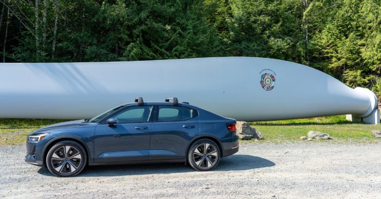 A dark blue car parked on gravel next to a huge metal wind turbine blade with pine trees behind.
