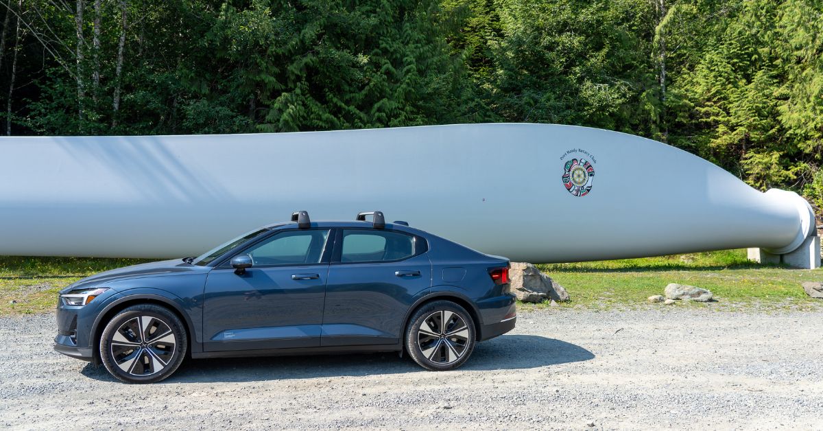 A dark blue car parked on gravel next to a huge metal wind turbine blade with pine trees behind.