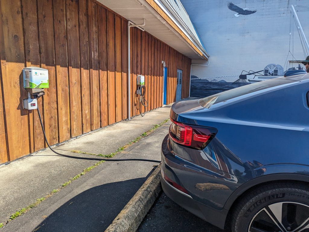 The back of a dark blue car parked next to a wooden wall with EV chargers.