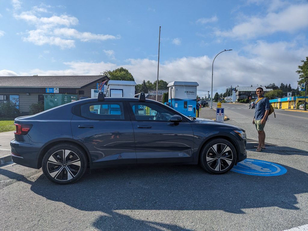 A man standing next to a dark blue car parked in an electric vehicle charging bay with buildings and a street in the background.