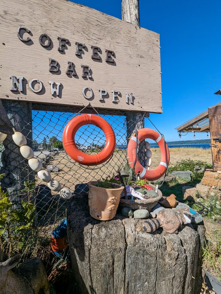 A wooden sign on a fence with upraised letters on it saying "Coffee bar now open." Small red life savers hang off the sign and plants and other items sit on a large tree stump below. In the background is brown grass, water and blue sky.