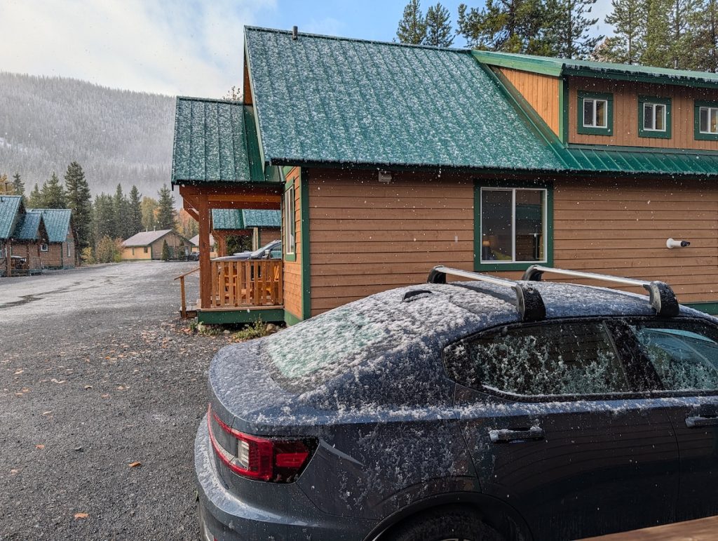 A car parked next to a cabin with more cabins and a hill with pine trees in the background, everything covered in a light layer of snow.