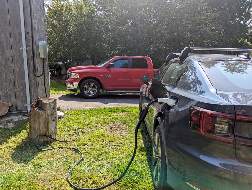 An electric vehicle plugged into a charger mounted on the wall of a wooden building, with a tree stump on the grass in between and a red Ram truck in the background.