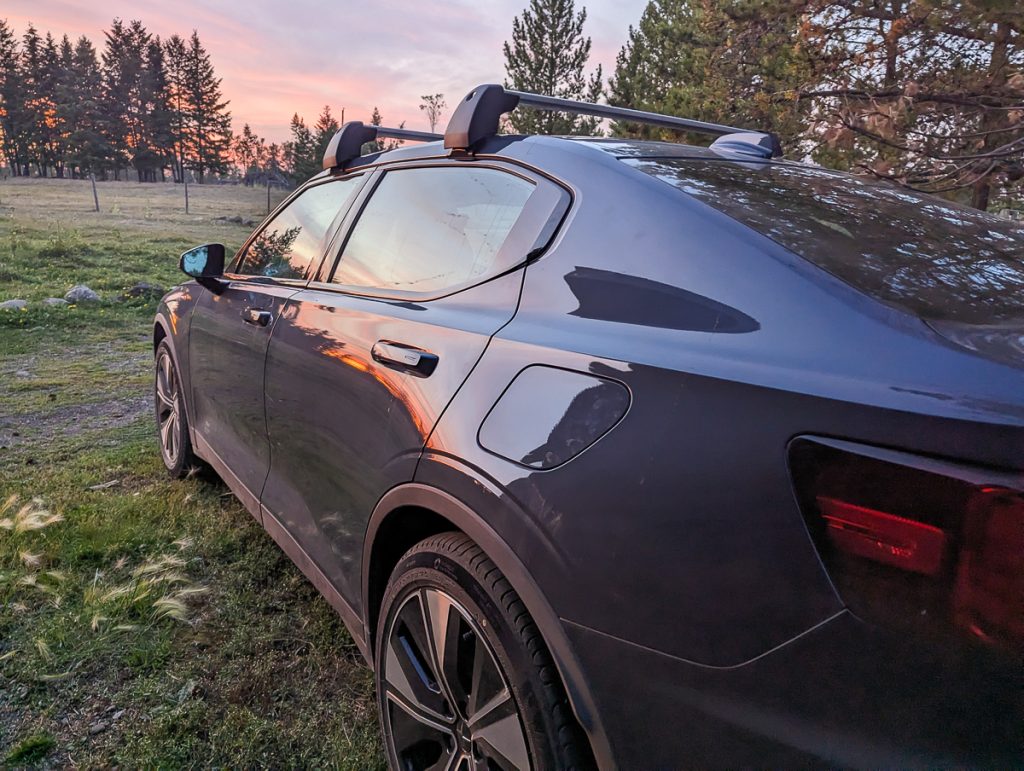 A dark blue car parked on grass with pine trees and a pink cloudy sunset in the background.