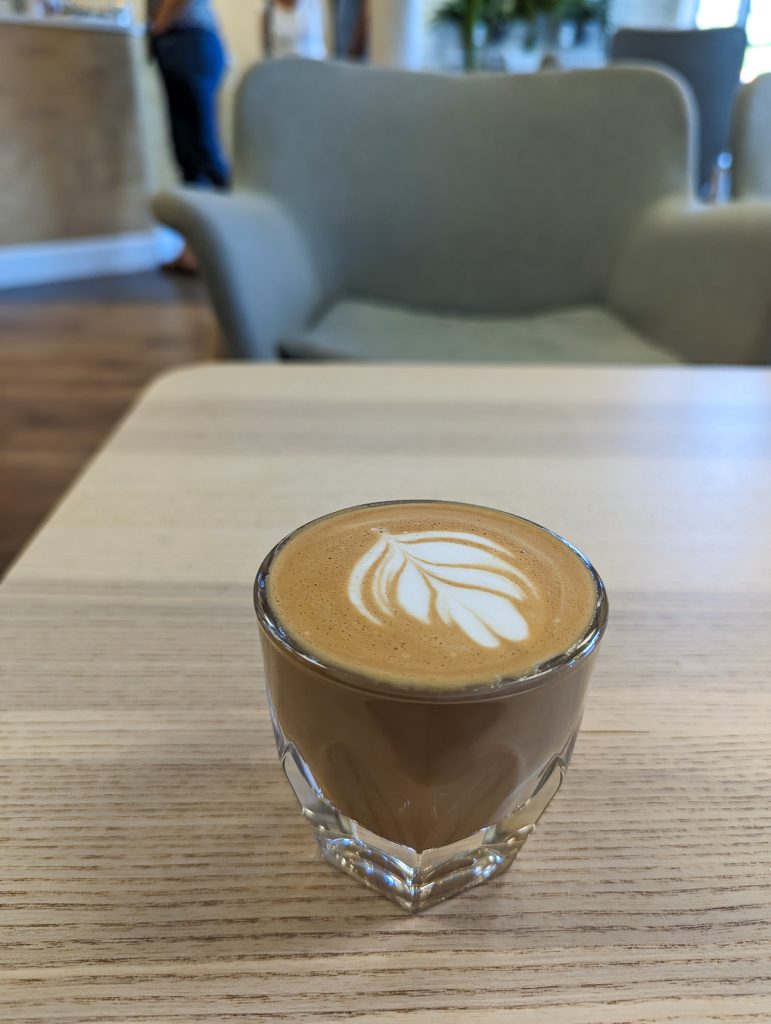 A close up of a coffee in a glass cup with a leaf drawn in the foam sitting on a wooden surface.