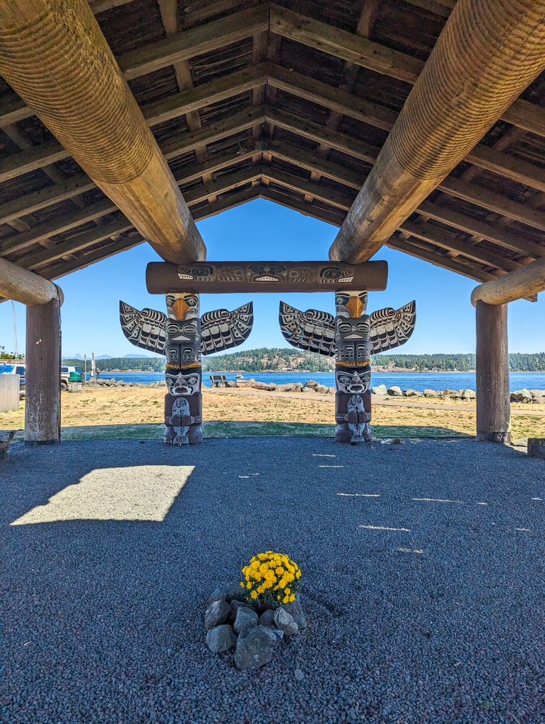 A small bunch of yellow flowers surrounded by large rocks on gravel with two carved wooden totem poles behind and a wooden ceiling above.