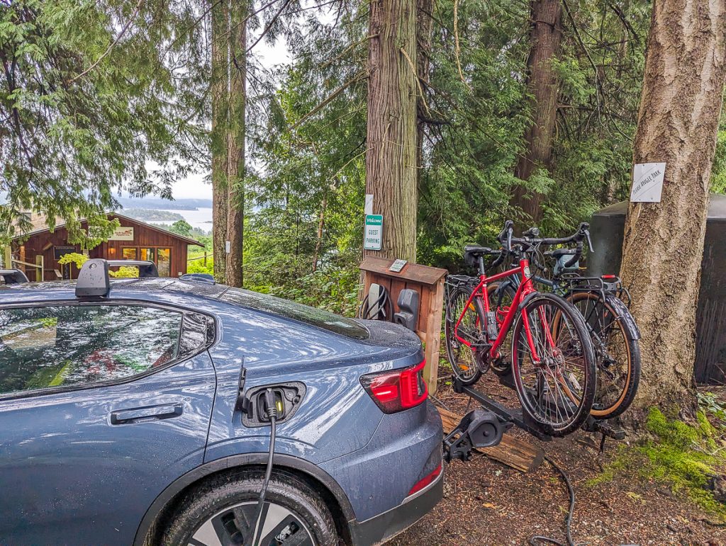 A photo of the end of an electric car plugged into a charger next to trees with two bikes on a tow-bar bicycle rack.