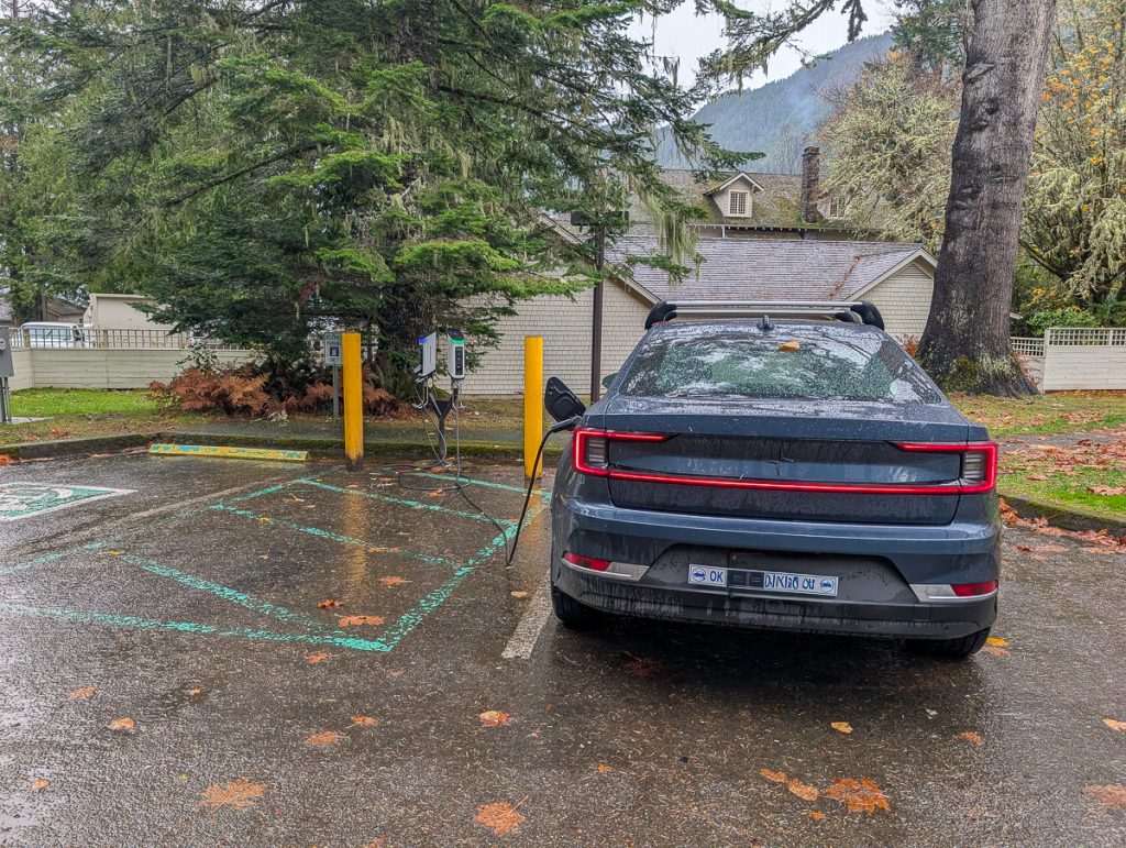 An electric vehicle plugged into a charging bay with another empty bay next to it and pine trees and a building in the background.