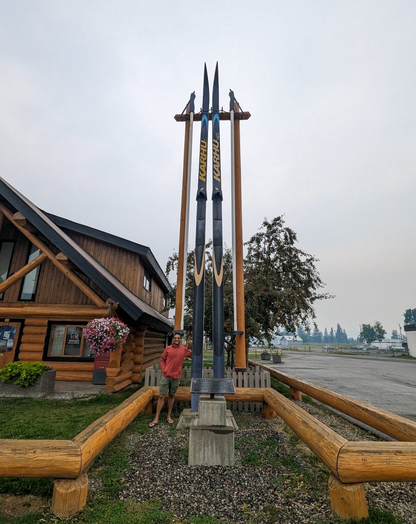 A man standing next to a huge sculpture of skis next to a wooden building on an overcast day.