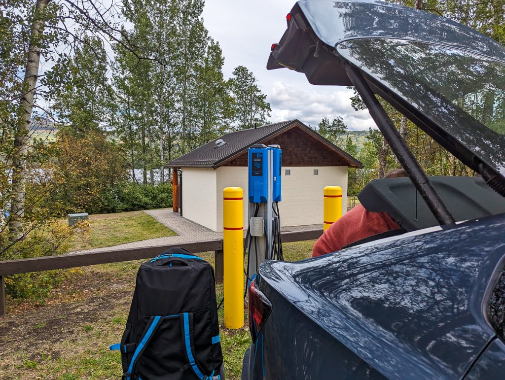 The open back end of a dark blue car with boot open plugged into an EV charger, with a backpack sitting on the ground next to it and a small building with trees in the background.