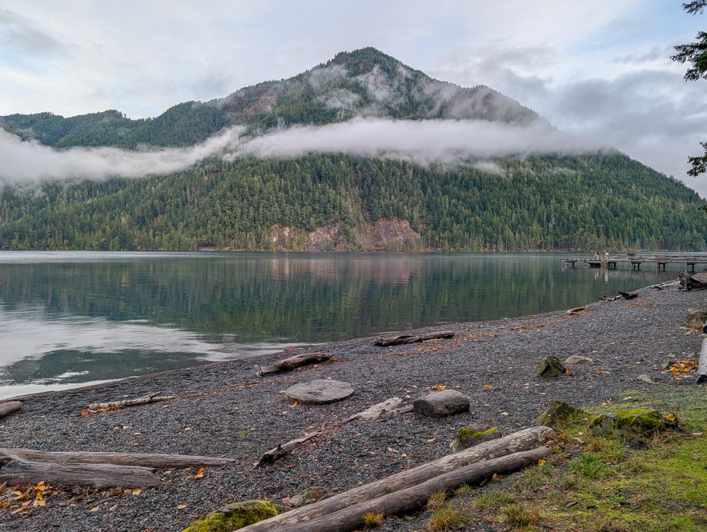A pebbled shore with driftwood, rocks and moss next to calm water with a mountain covered in pine trees and misty clouds in the background.